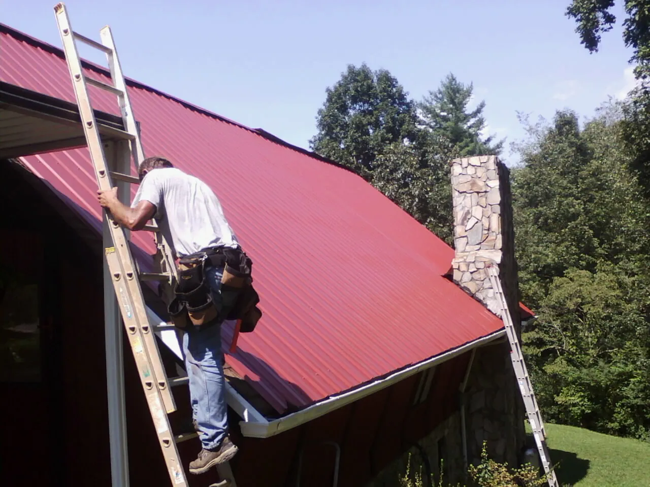 Roofer on a ladder beside a red metal roof and stone chimney — Soffit & Fascia Repair by Roofing Brothers in Grafton